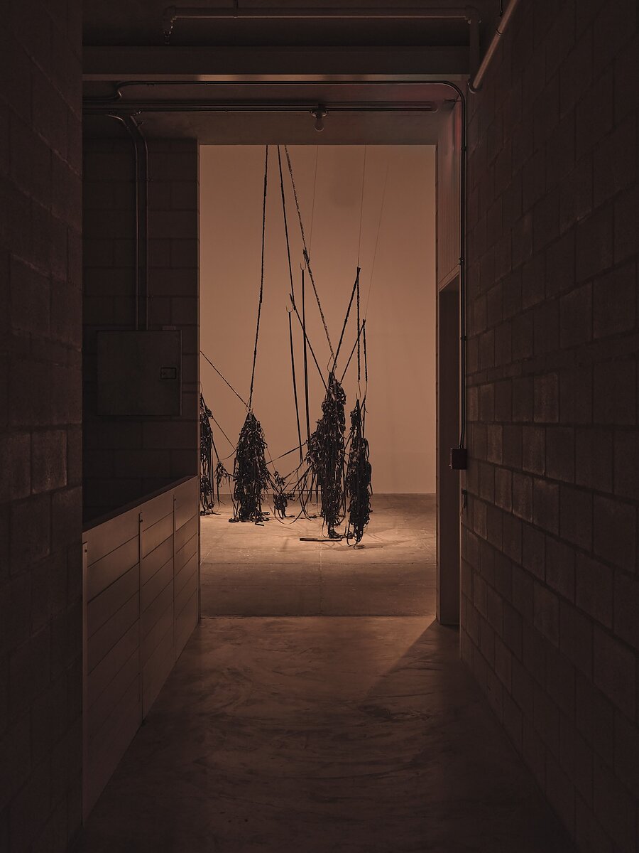 Partial view from a hallway looking onto a work of art composed of tangled black fabric hanging from the rafters precariously by sharp hooks.