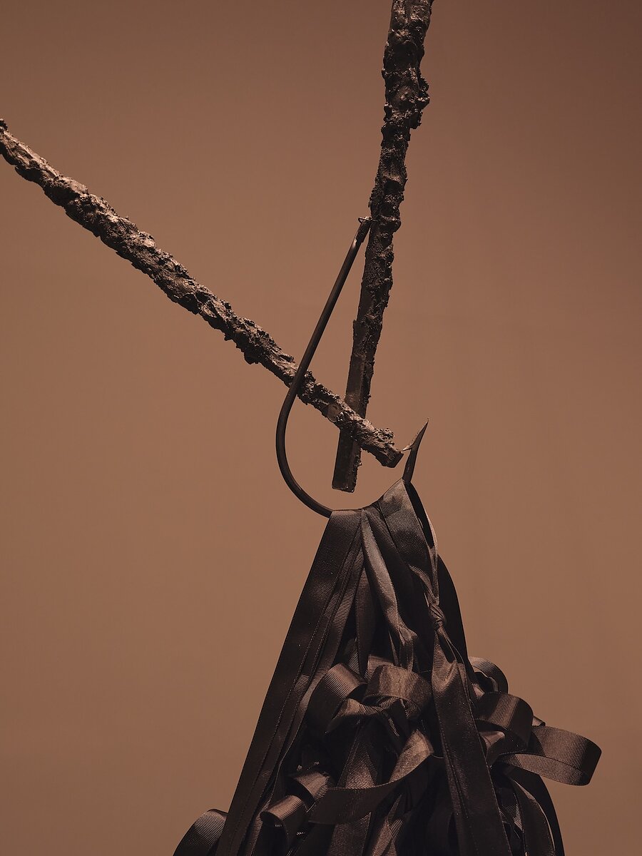 A close up of a work of art featuring a tangle of shiny black ribbons of fabric suspended by a large hook and crusted-over rods.