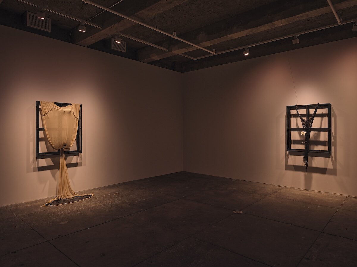 A dimly lit gallery featuring two works hanging on the walls, each featuring draped fabric hanging heavily from black, frame-like structures.